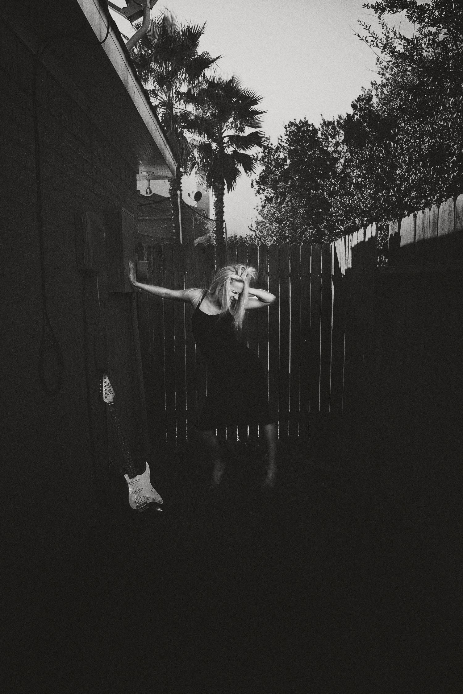 Black and white photo of a woman in a dark dress posing dramatically against a wooden fence with palm trees behind her.