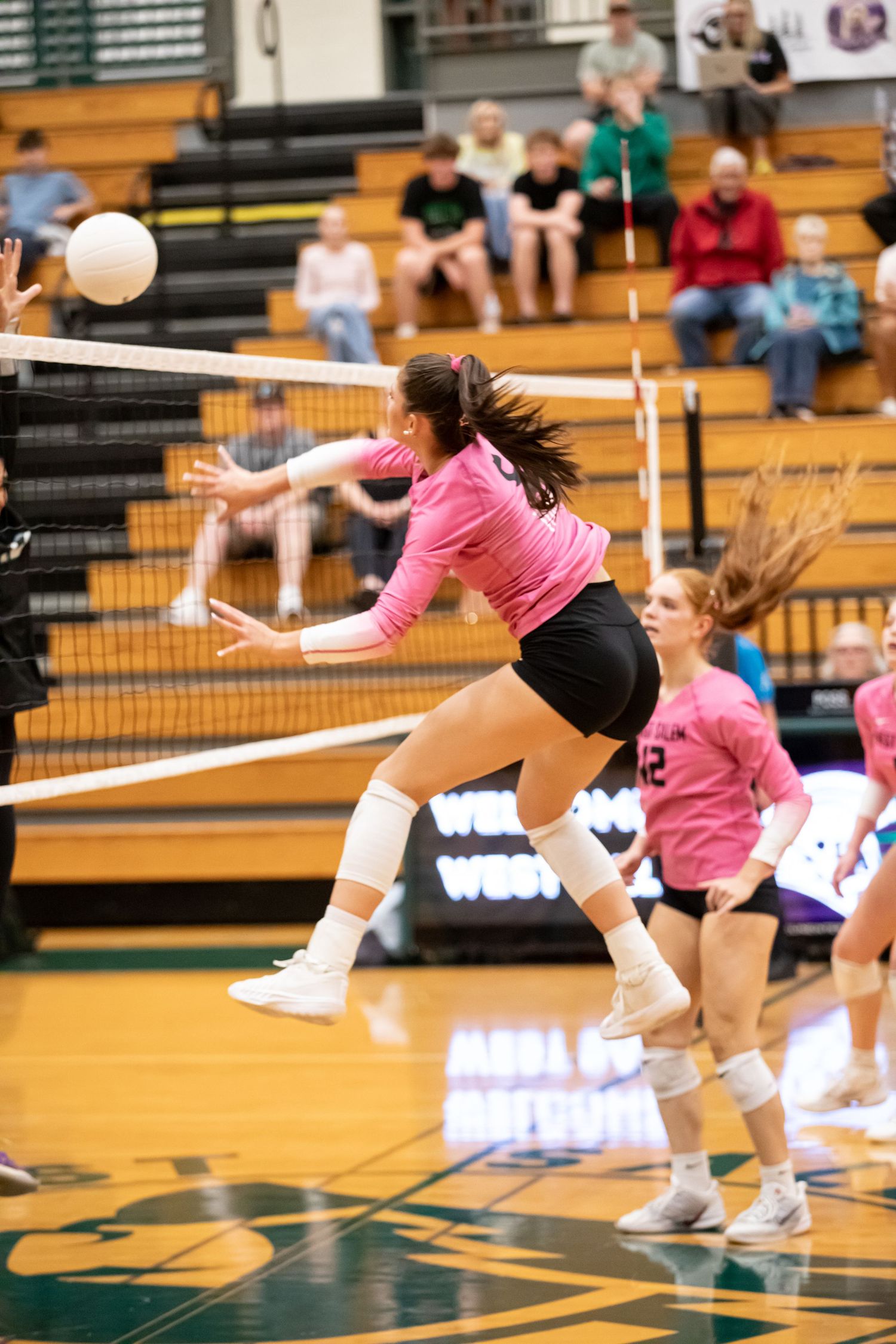 Female volleyball player in a pink jersey leaps toward the net to hit the ball during a high school gym match as spectators watch.
