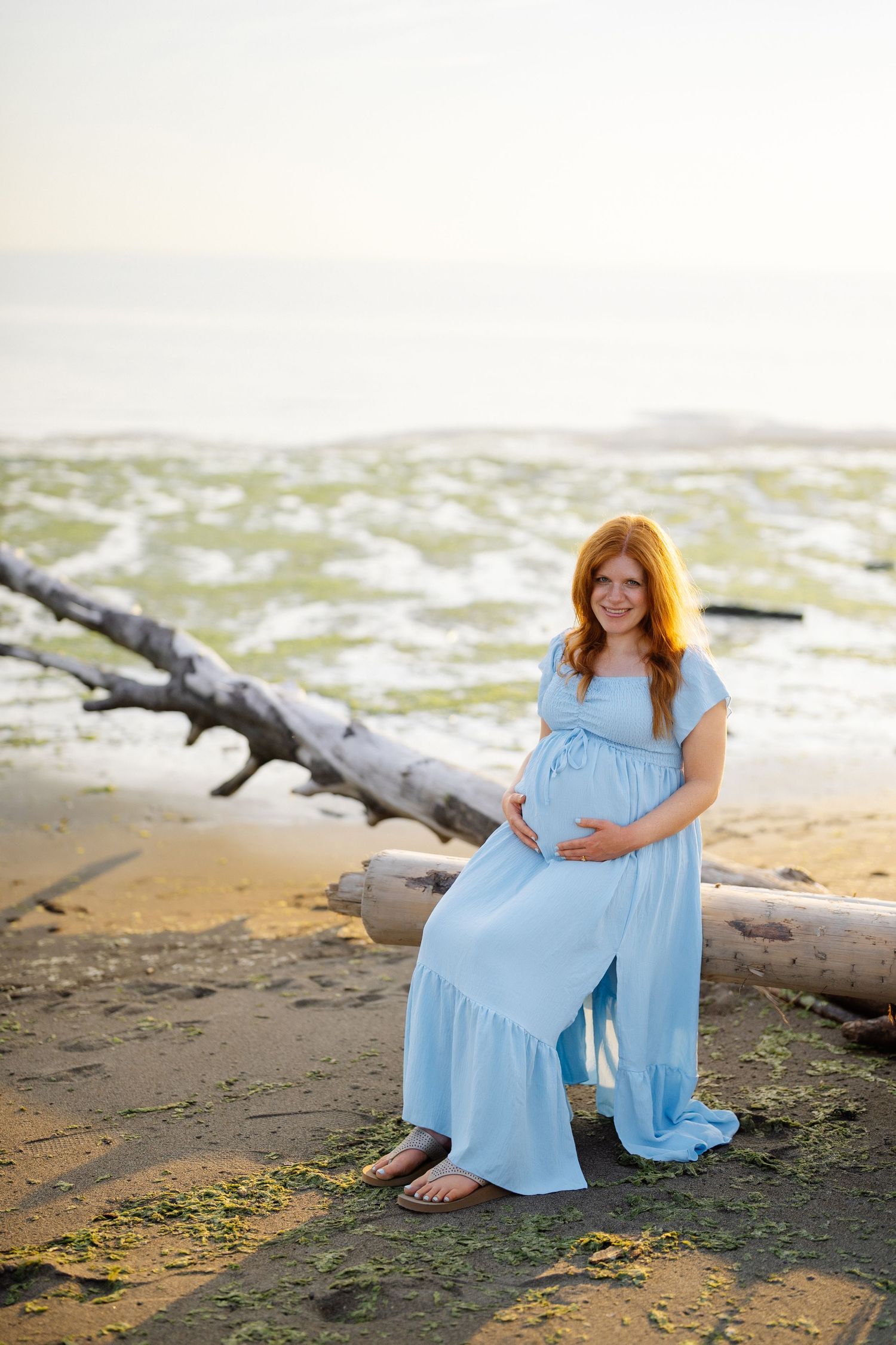 Pregnant woman in a flowing light blue dress sits on driftwood, cradling her baby bump on a sandy beach at sunset.