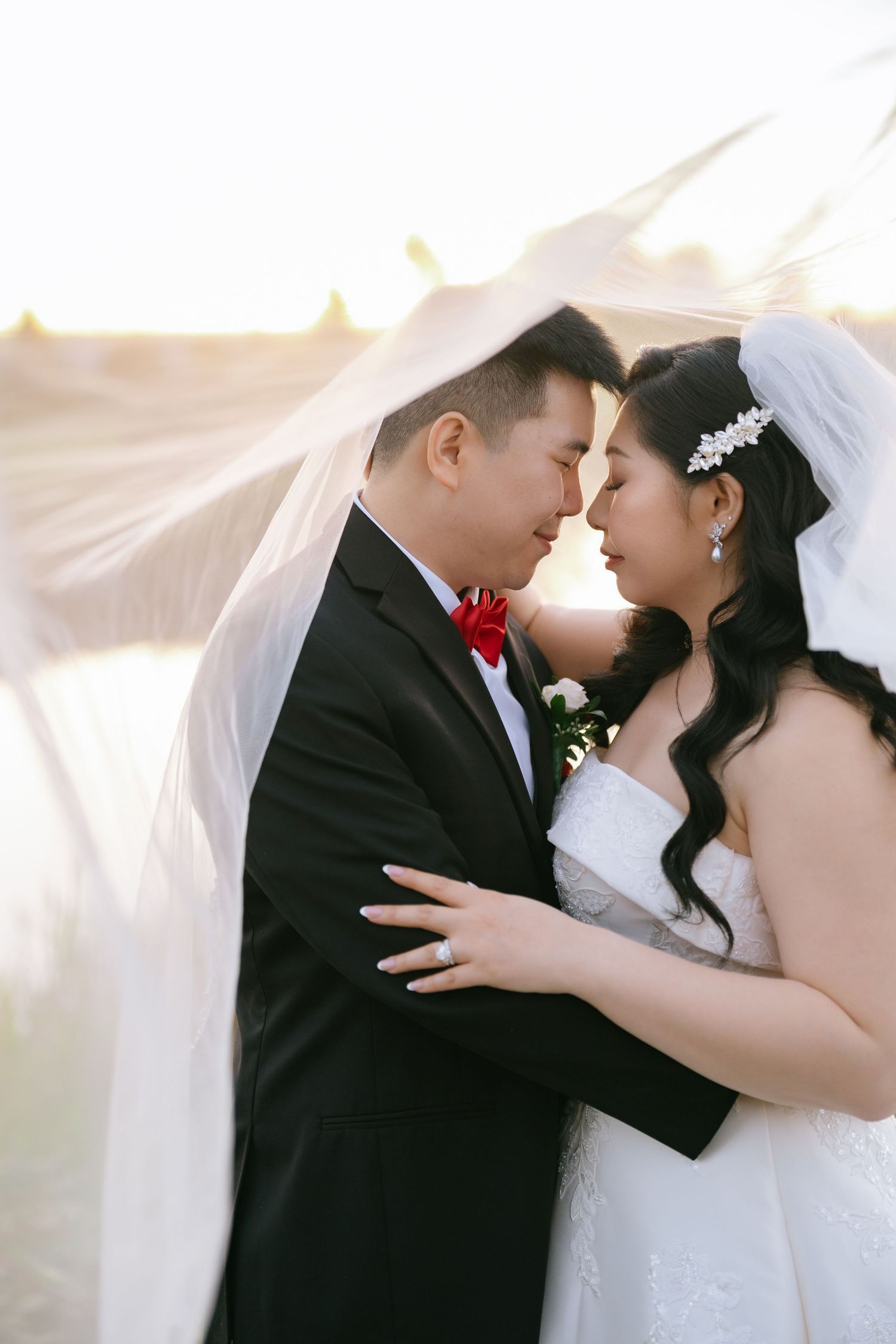 Bride and groom share an intimate moment forehead to forehead beneath a flowing sheer veil at sunset, she in a white gown, he in a tuxedo.