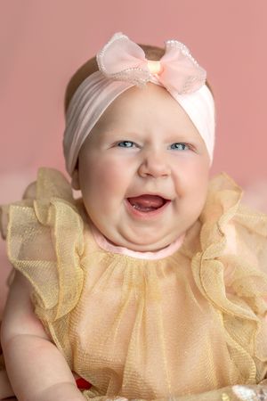 Smiling baby girl in a golden ruffled dress and pink bow headband poses against a soft pink background.