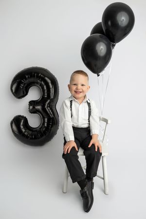 Smiling boy celebrating his 3rd birthday, sitting on a chair with black balloons and a number 3 balloon on white background.