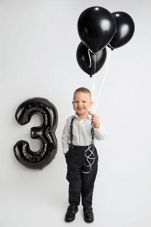 Smiling boy in suspenders holding black balloons beside a large number 3 balloon, celebrating his 3rd birthday.