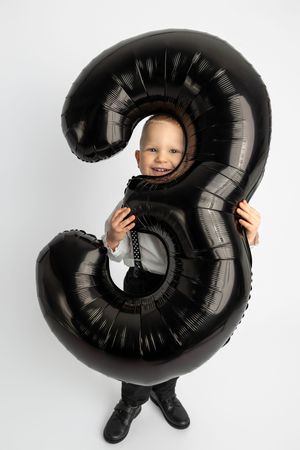 Smiling young boy peering through a large black number 3 balloon on white background, perfect for 3rd birthday celebration.