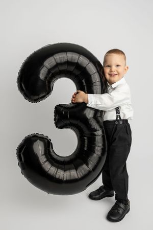 Smiling boy in white shirt and suspenders holding large black number 3 balloon on white background.
