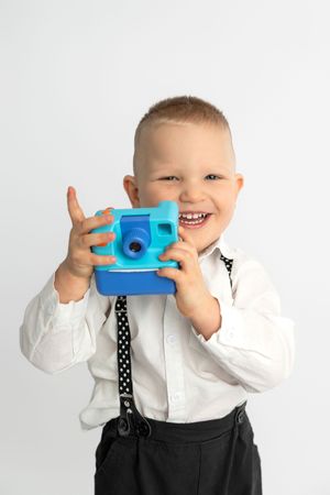 Smiling young boy in white shirt and suspenders holding a blue toy camera up to take a photo.