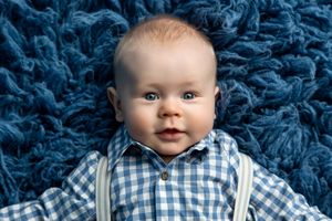 Adorable baby boy with blue eyes lying on blue fluffy carpet wearing checkered shirt with suspenders.