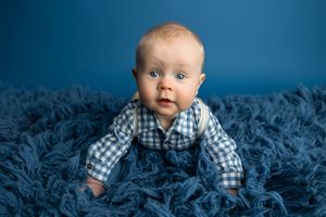Adorable baby boy with blue eyes in plaid shirt sits on fluffy blue rug against blue background.
