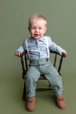 Smiling baby boy in blue shirt, suspenders, and green pants sitting on a wooden chair against an olive green background.