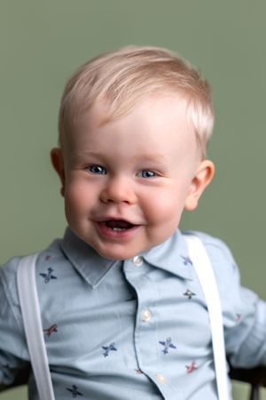 Smiling blonde toddler boy with blue eyes wearing a light blue button-up shirt with suspenders against a green background.