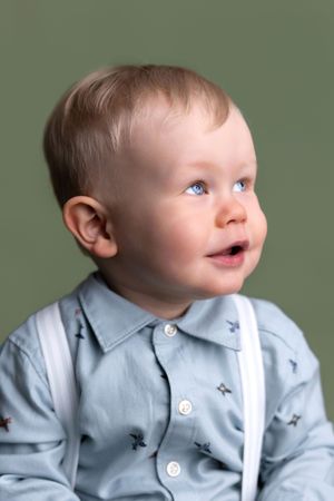 Adorable blue-eyed baby boy in a light blue star-print shirt with white suspenders, looking up against green background.
