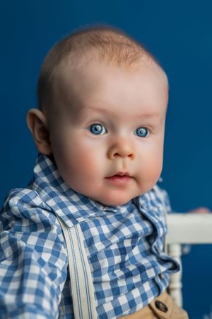 Adorable baby with blue eyes wearing a blue gingham shirt and suspenders, sitting against a blue background.