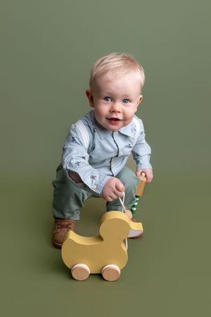 Smiling baby boy with blue eyes playing with a wooden duck pull toy, wearing a light blue outfit on a green background.