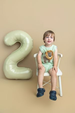 Smiling toddler in mint green outfit sits on white chair beside number 2 balloon, celebrating 2nd birthday.