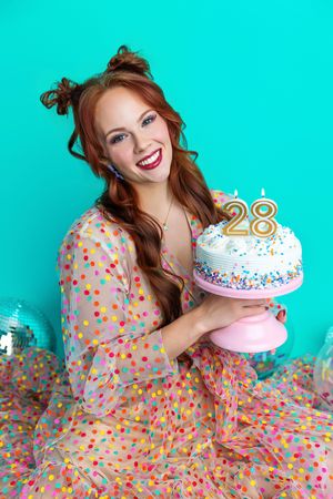 Smiling redhead woman in polka dot dress holding a white birthday cake with number 28 candles on teal background.