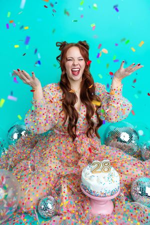 A joyful redhead woman in a polka dot dress celebrates her 28th birthday with confetti, disco balls, and a cake.