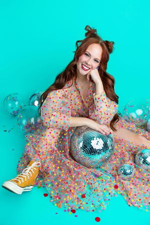 Smiling redhead woman in colorful polka dot dress sitting with disco balls and confetti on teal background.
