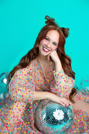 Smiling redhead woman in colorful polka dot dress posing with a disco ball on a turquoise background.
