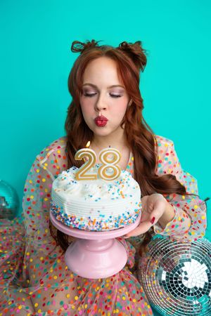 Young woman with red hair blowing out candles on a white birthday cake with number 28, against teal background.