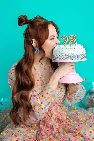 Young woman in polka dot dress biting into a white birthday cake with number 28 candles on teal background.