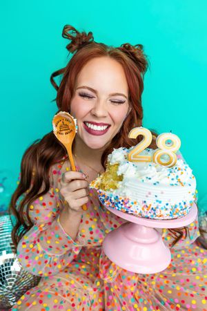 Smiling redhead woman holding wooden spoon beside a colorful sprinkle birthday cake with number 28 candles on pink stand.