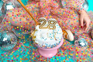 A festive 28th birthday cupcake with white frosting and sprinkles on a pink stand, surrounded by disco balls and confetti.
