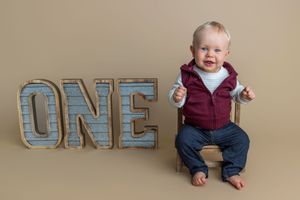 A cheerful baby boy sits on a wooden box next to large ONE letters, celebrating his first birthday.