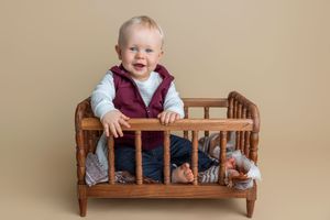Smiling baby boy in maroon vest standing in a small wooden toy crib against a beige studio background.