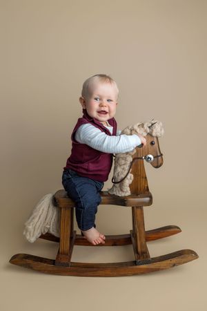Smiling baby boy in burgundy vest sitting on wooden rocking horse toy against beige background.