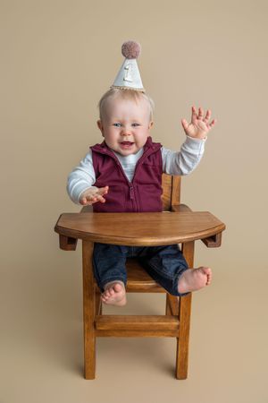 A happy baby wearing a birthday hat sits in a wooden high chair, raising hands joyfully, dressed in a maroon vest and jeans.