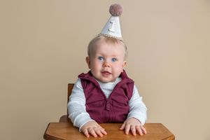 Adorable blue-eyed baby wearing a birthday party hat sits at a wooden high chair against a beige background.