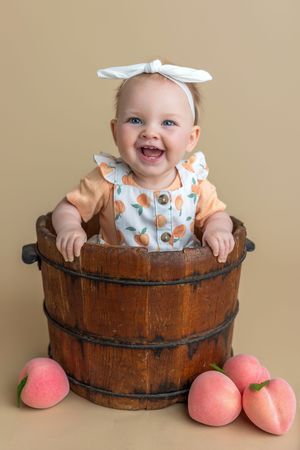 Smiling baby girl in peach outfit sitting in rustic wooden bucket surrounded by fresh peaches.