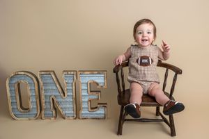 A smiling baby in a brown romper sits on a wooden chair next to large ONE letters for a first birthday photo.