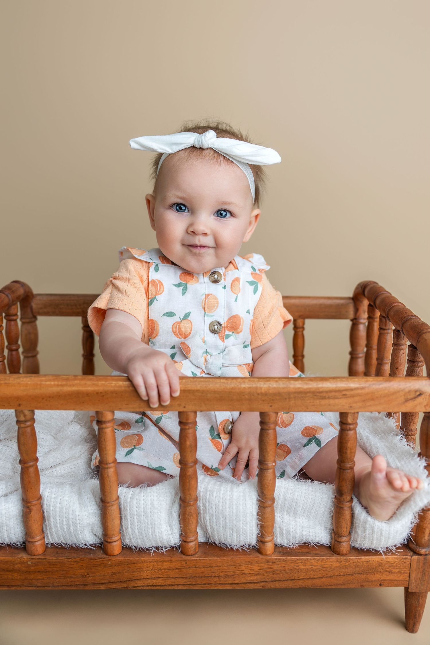 Smiling baby girl with blue eyes and white bow headband sitting in a wooden crib wearing a peach-print romper.