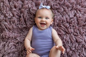 Smiling baby girl in lavender onesie with bow headband lying on fluffy mauve carpet, looking up happily.