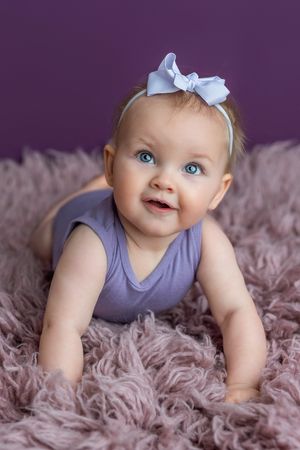 Adorable baby girl with blue eyes and bow headband smiles while doing tummy time on a fluffy purple rug.