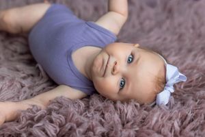 A cute baby in a purple onesie with a white bow lies on a fluffy mauve rug, gazing up with bright blue eyes.