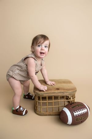 A cute baby in a checkered romper stands by a wicker basket next to a Wilson football on a beige background.