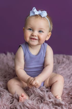 Smiling baby girl in lavender onesie and bow headband sitting on pink fur rug against purple background.