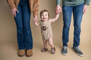 A baby in a football romper holds hands with parents while learning to walk against a neutral beige background.