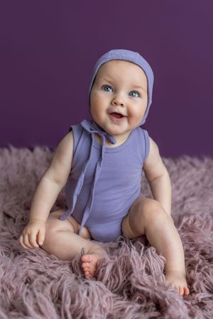 Smiling baby in purple bonnet and bodysuit sitting on fluffy lavender rug against purple background.
