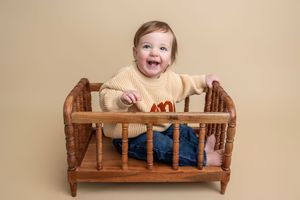 Smiling baby in yellow sweater and jeans sitting in a small wooden crib against a beige background.