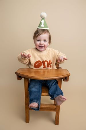 Smiling baby in a green number 1 party hat and football sweater sits on a wooden chair celebrating their first birthday.