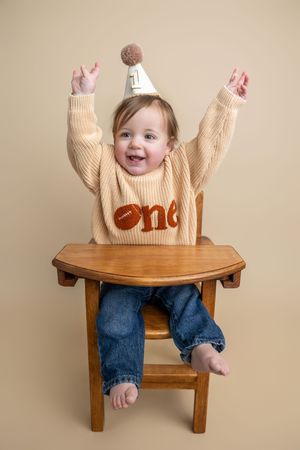 Baby's first birthday portrait in high chair, wearing “one” sweater and party hat, arms raised joyfully.