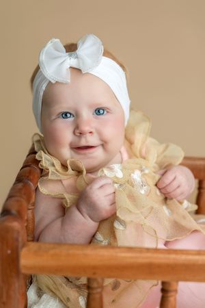 Smiling baby girl with blue eyes wearing a gold ruffle dress and white bow headband sitting in a wooden chair.