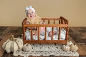Smiling baby in white bow sits in wooden doll crib surrounded by white pumpkins on rustic wood floor.