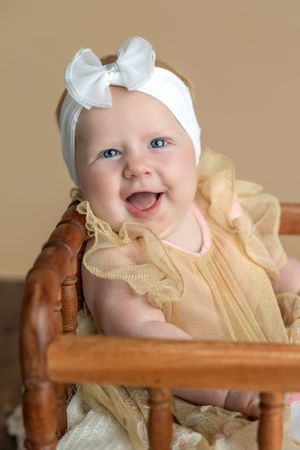 Smiling baby girl with blue eyes wearing a yellow dress and white bow headband sitting in a wooden chair.