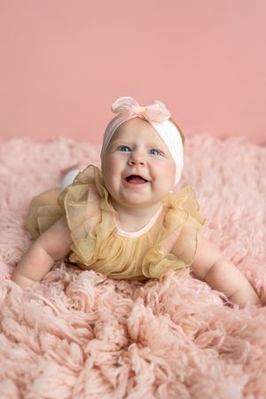 Smiling baby girl in yellow ruffle dress and pink bow headband lying on fluffy pink blanket against pink background.