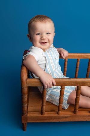 Smiling baby boy in striped romper sitting in a wooden mini crib against a blue background.