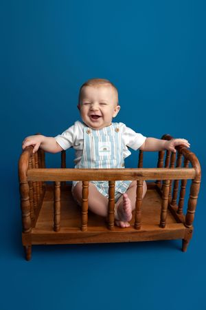 Happy baby in striped romper laughing in a wooden crib against a blue background.